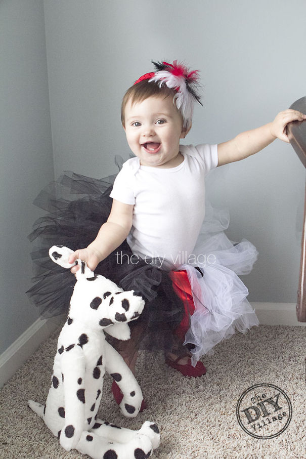 Toddler wearing a black white and red tutu with a feather headband holding a stuffed spotted dog for children's Halloween costume ideas.