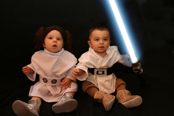Two toddlers dressed in themed children's Halloween costume ideas, one holding a glowing lightsaber toy on black background.