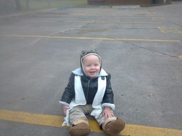 Toddler smiling while sitting on pavement dressed in a creative children's Halloween costume idea with aviator hat and boots.