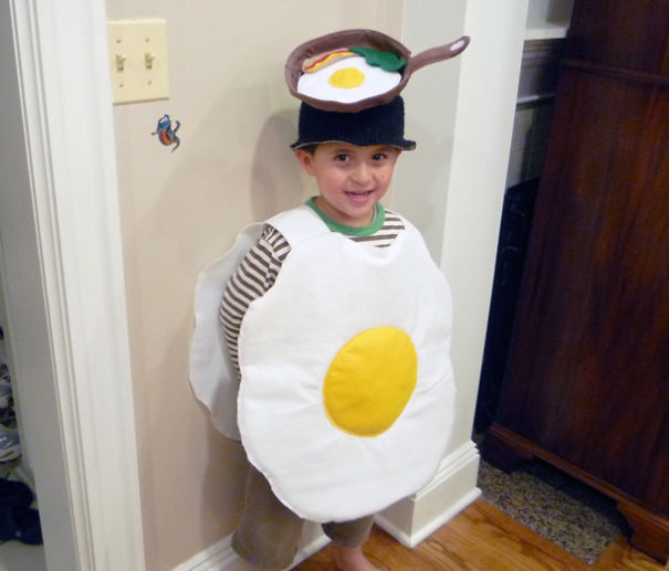 Child wearing a creative children's Halloween costume idea dressed as a fried egg with a skillet hat indoors.