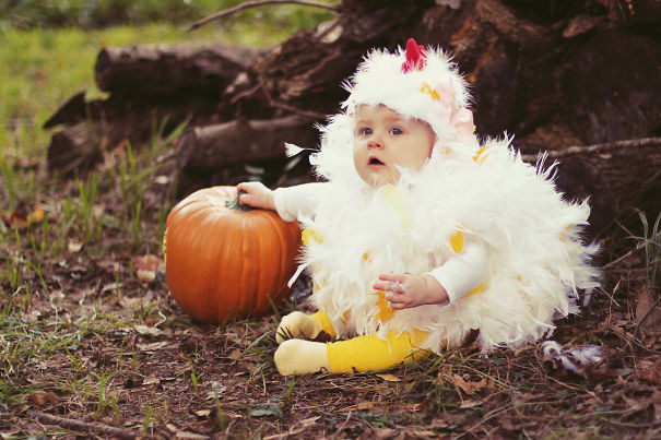 Baby in a fluffy chicken costume sitting outdoors next to a pumpkin in children's Halloween costume ideas setting