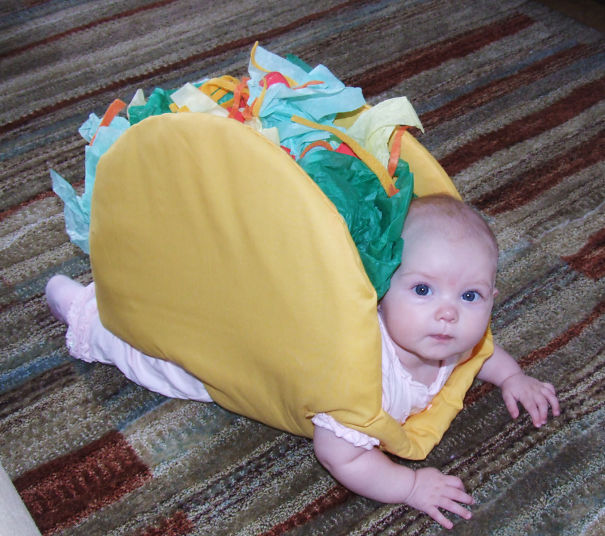 Baby dressed in a creative children's Halloween costume crawling on a carpet in a taco outfit.