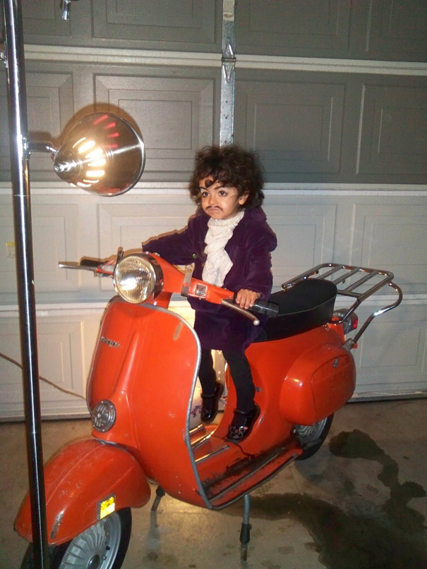 Child in creative children's Halloween costume ideas outfit sitting on a vintage orange scooter in a garage.