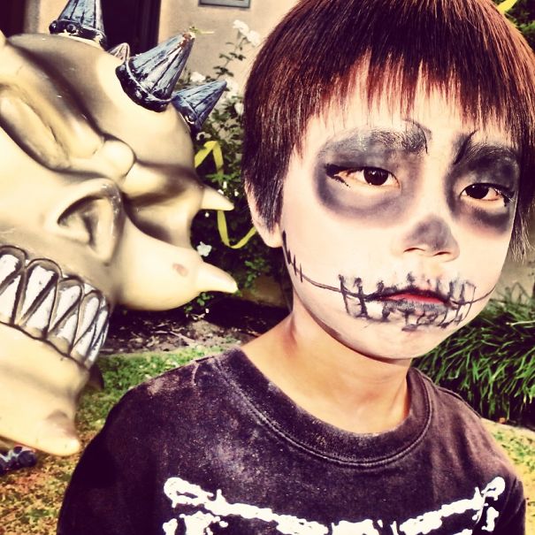 Child with spooky Halloween face paint and costume, posing next to a large monster prop outdoors for children's Halloween costume ideas.