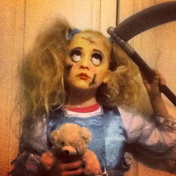Child in creepy doll Halloween costume holding a teddy bear and a large scythe for children's Halloween costume ideas.