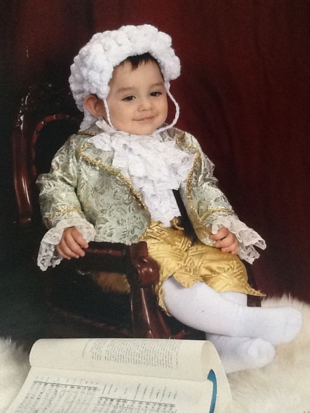 Toddler dressed in an ornate historical costume sitting on a chair, a book open nearby, children's Halloween costume ideas inspiration.
