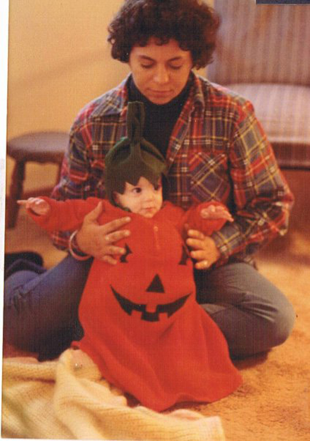 Baby dressed in a pumpkin costume sitting on adult’s lap indoors, a cute example of children's Halloween costume ideas.