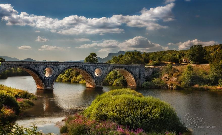 Ancient mystical stone bridge over calm river with lush greenery and dramatic cloudy sky in a serene natural setting.
