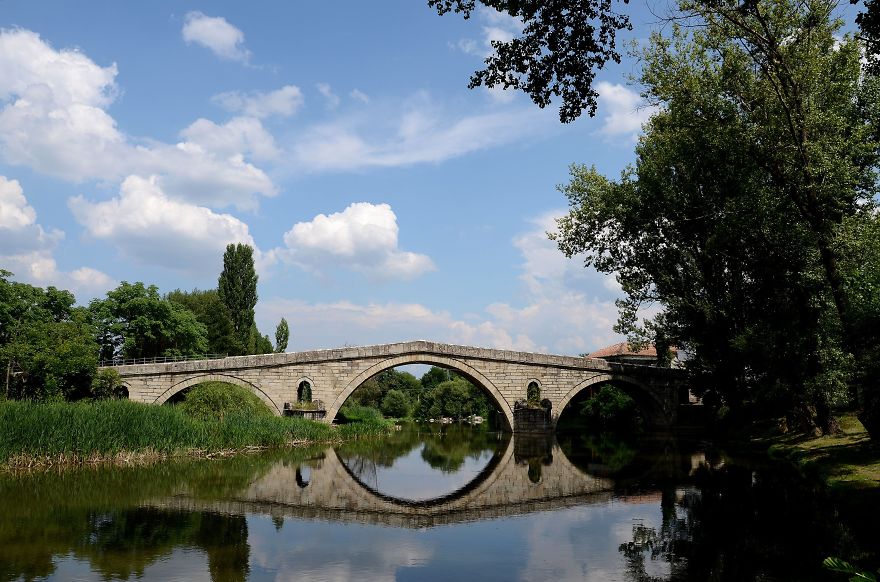 Ancient stone mystical bridge over calm river surrounded by lush greenery under a partly cloudy sky.