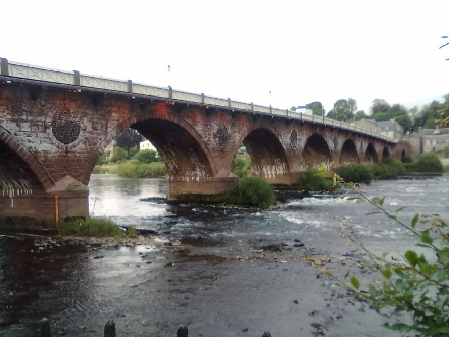 Ancient mystical stone bridge with multiple arches spanning over a flowing river surrounded by greenery.
