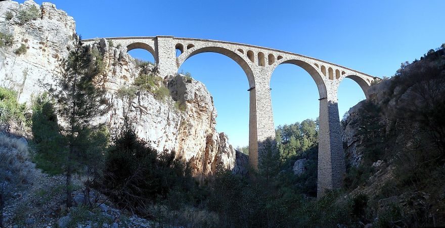 Stone mystical bridge with large arches spanning a rocky gorge under clear blue sky in a natural landscape.