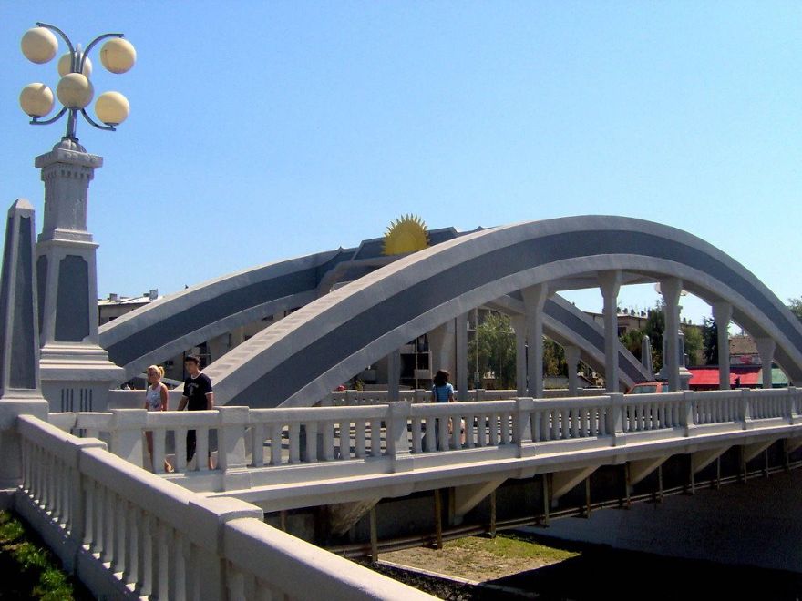 Arched mystical bridge with pedestrians, featuring art deco design and bright lamps under clear blue sky.