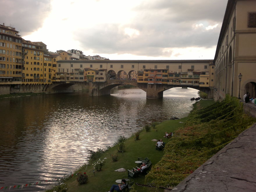Historic mystical bridge over calm river with colorful buildings and people relaxing on nearby grassy bank.