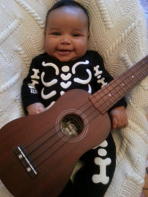 Smiling baby in a skeleton Halloween costume lying on a blanket while holding a wooden guitar for children’s costume ideas.