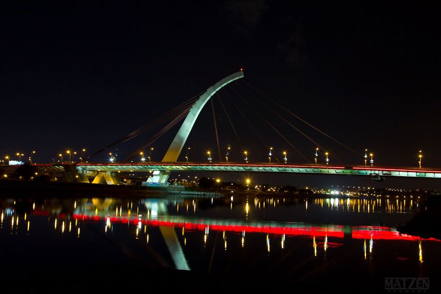 Illuminated mystical bridge at night with colorful lights reflecting on calm water in a serene setting.