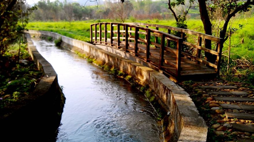 Wooden mystical bridge over a narrow flowing stream surrounded by green fields and trees on a sunny day.