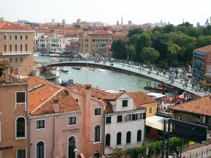 Glass pedestrian bridge over a canal in a historic city, with people walking and old buildings nearby, showcasing mystical bridges.