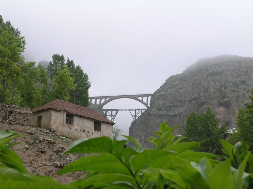 Arch bridge rising through misty mountains near rustic house surrounded by lush greenery and foggy landscape, mystical bridges scene