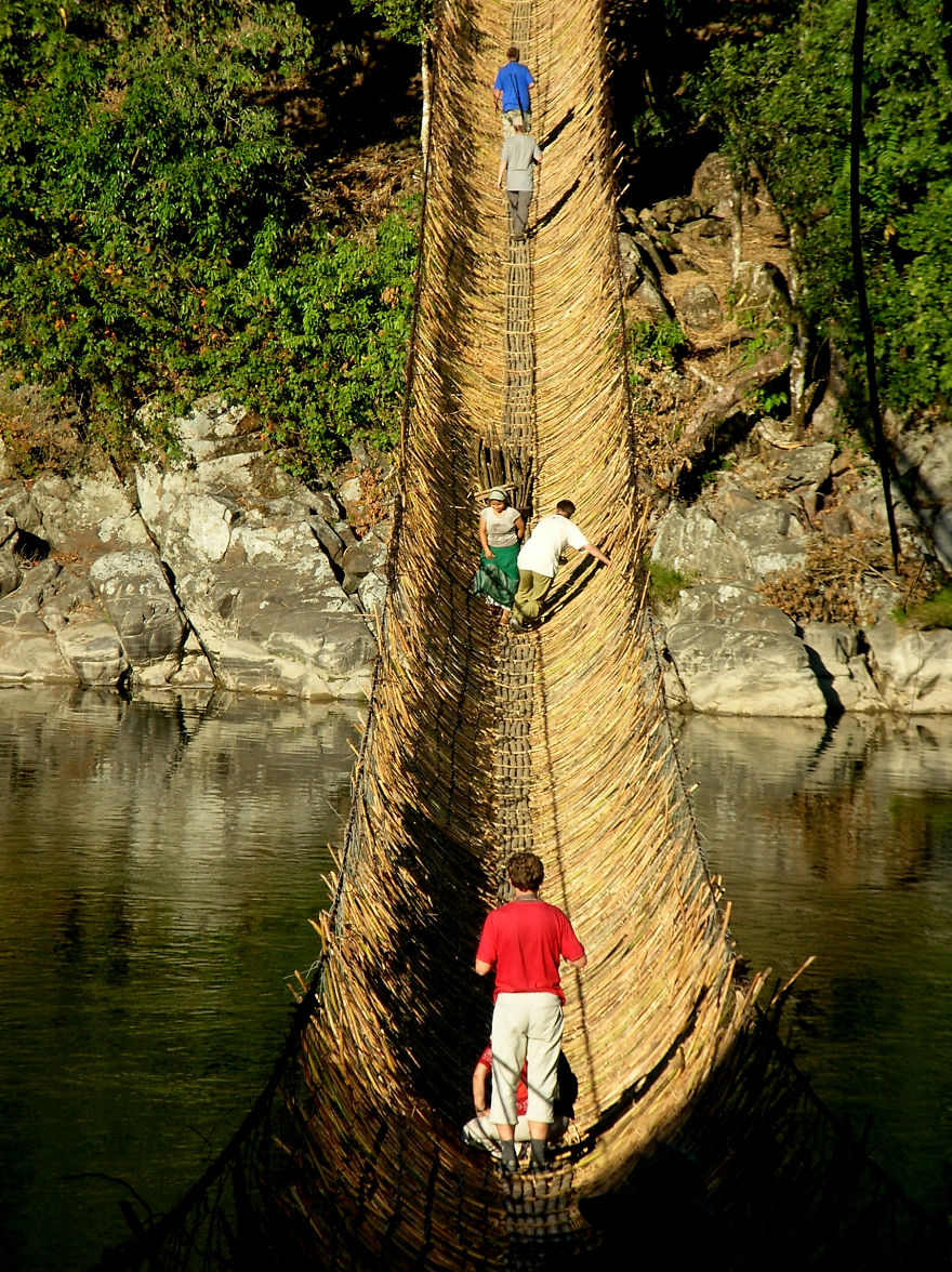 Handcrafted mystical bridge made of woven vines spanning a river with people walking across surrounded by nature.