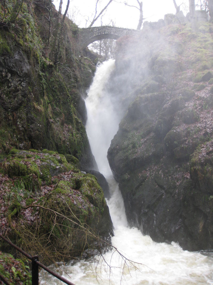 Stone mystical bridge over a powerful waterfall surrounded by moss-covered rocks and mist in a forest setting