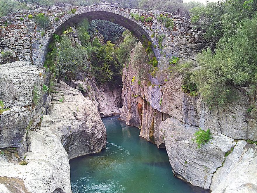 Ancient mystical stone bridge arching over a turquoise river flowing through a rocky canyon surrounded by greenery.