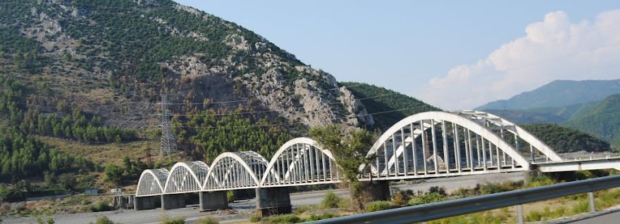 Long white mystical bridge with arches spanning a river, set against a backdrop of forested hills and blue sky.