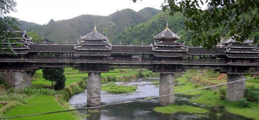Ancient wooden mystical bridge with traditional architecture spanning a river in a lush green mountainous landscape.