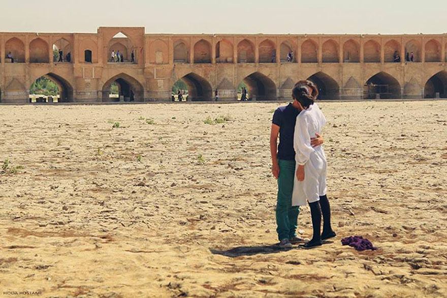 Couple standing on dry riverbed near a mystical bridge with multiple arches under a clear sky.