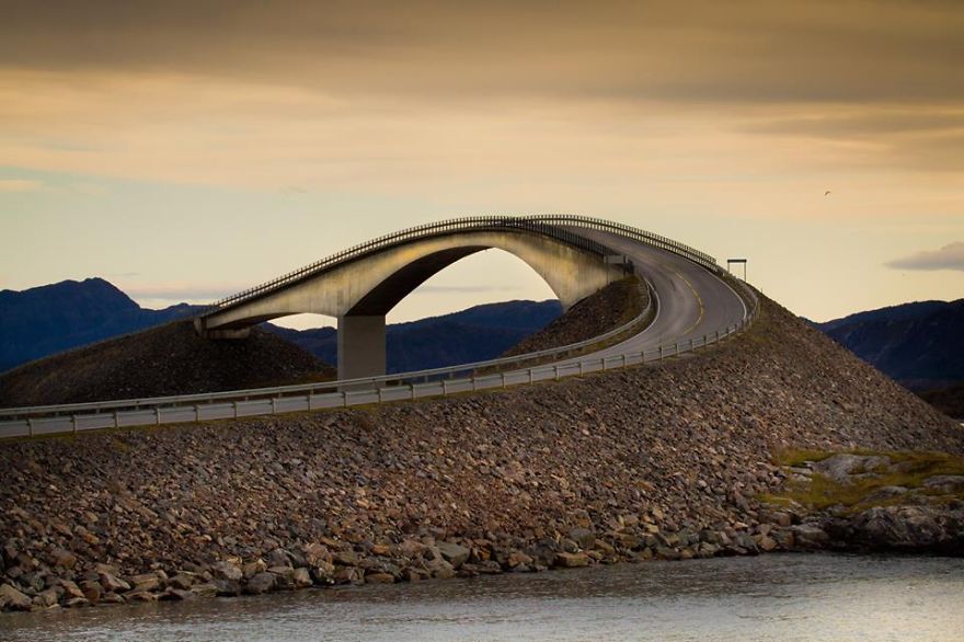 Curved mystical bridge over rocky terrain at sunset with mountains in the background, evoking a sense of otherworldly travel.