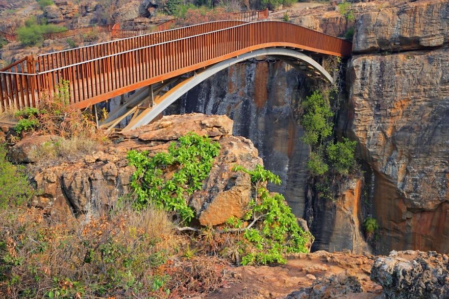 Rust-colored mystical bridge arching over a rocky canyon with sparse greenery, blending nature and architecture seamlessly.