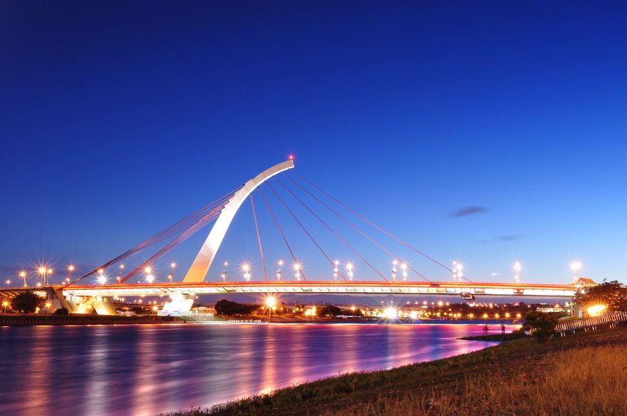 Mystical bridge illuminated at night over calm water with a clear deep blue sky in the background.