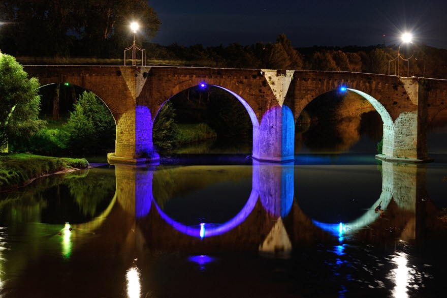 Stone bridge with mystical blue lighting reflecting on calm water at night, showcasing a mystical bridge scene.