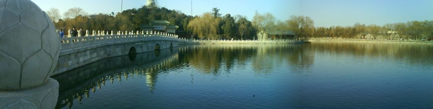 Stone mystical bridge with decorative railings spanning calm water surrounded by trees under a clear sky.