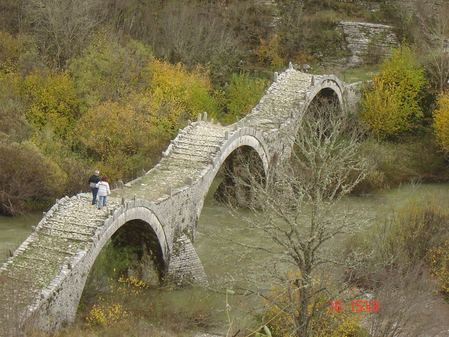 Ancient mystical stone bridge with multiple arches surrounded by autumn trees and flowing river below, two people walking across.