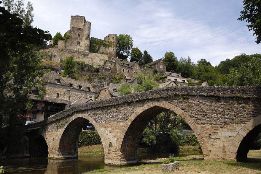 Ancient mystical stone bridge with arches over river, leading to old village and castle surrounded by greenery.