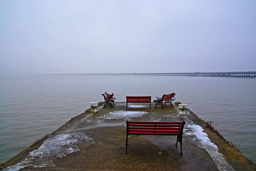 Empty red benches on a misty pier near a long mystical bridge fading into the fog over calm water.