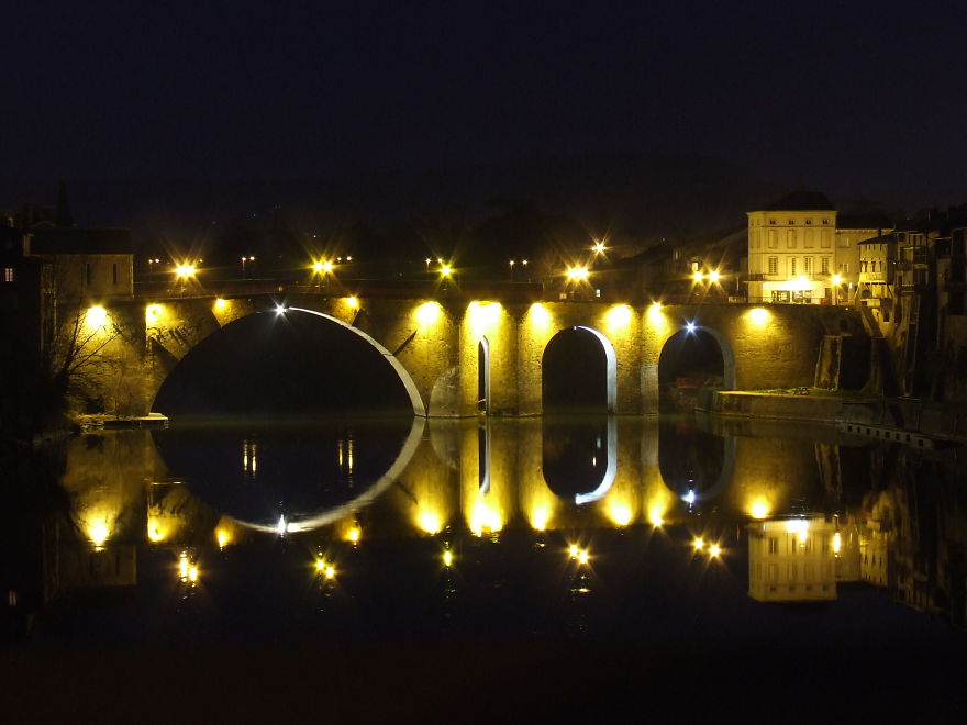 Ancient mystical bridge illuminated at night with golden lights reflecting on calm water, creating an enchanting scene.