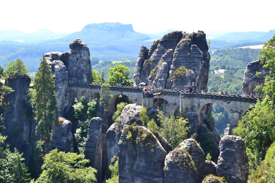 Ancient mystical stone bridge connecting towering rock formations surrounded by lush green forest under a bright sky.