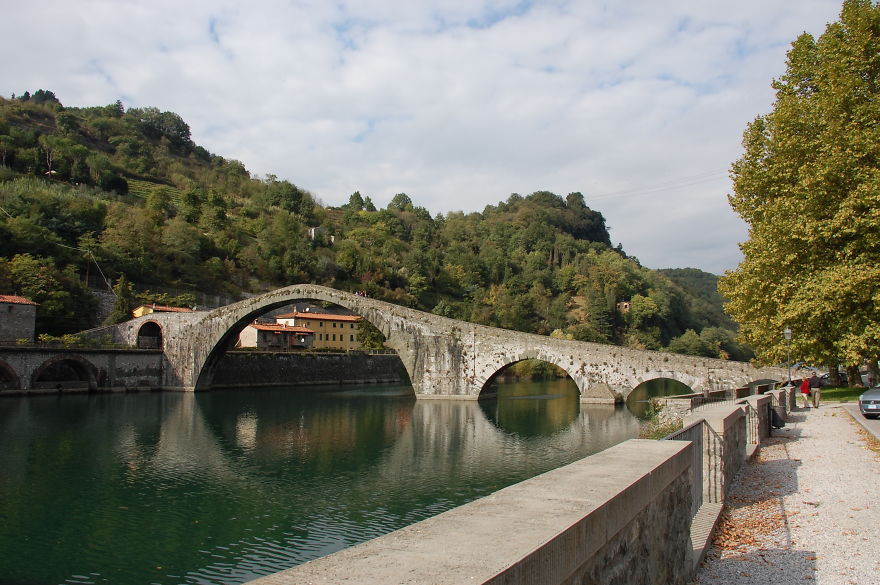 Ancient mystical stone bridge arching over calm river surrounded by lush green hills and autumn trees.