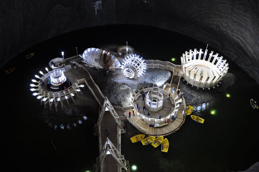 Mystical bridges and illuminated walkways over water inside a large dark cave with boats nearby at night.