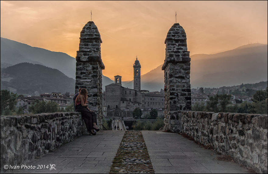 Mystical stone bridge at sunset with two people sitting, overlooking an ancient town and distant mountains.