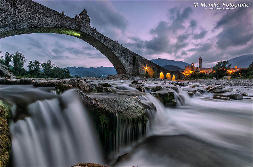 Mystical stone bridge arching over flowing river with soft waterfall and village lights at twilight in a scenic landscape.