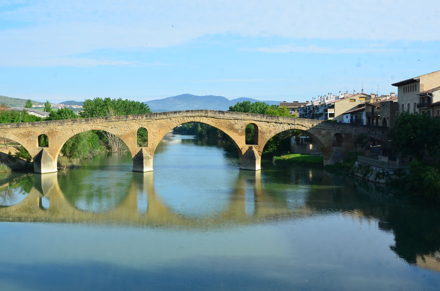 Ancient mystical stone bridge with multiple arches spanning a calm river under blue sky and distant hills.