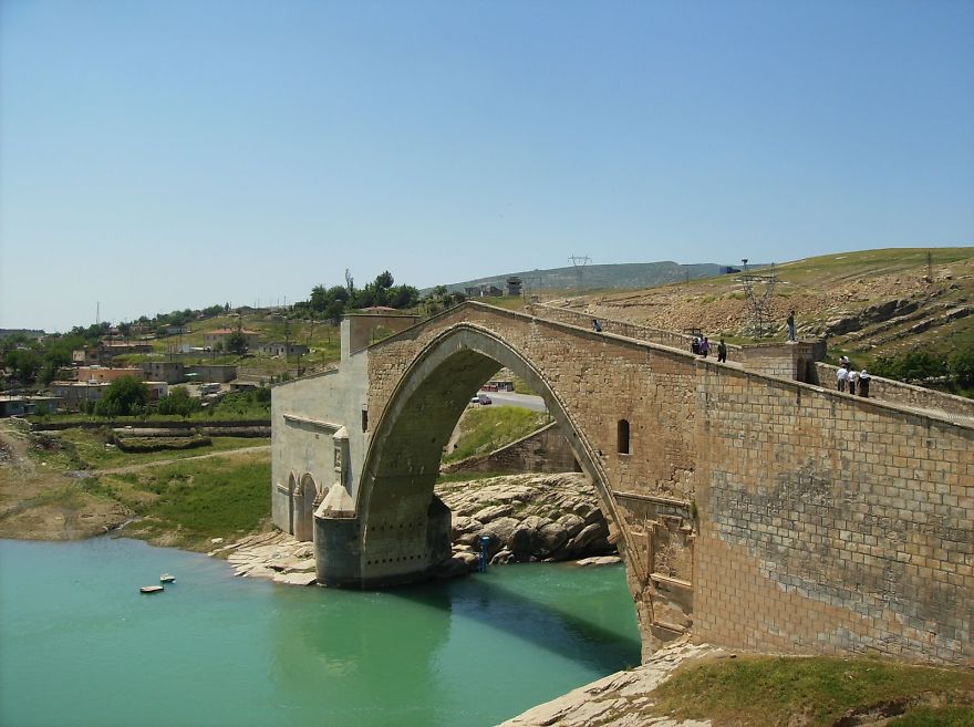 Ancient stone mystical bridge arching over turquoise water with people walking, set in a rural landscape under clear sky.