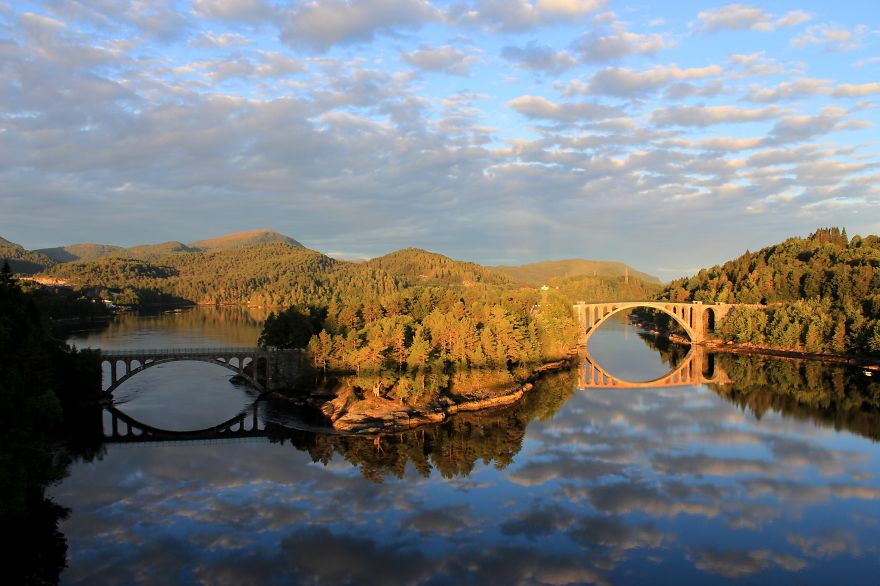 Two mystical bridges arching over calm water surrounded by forested hills under a partly cloudy sky.