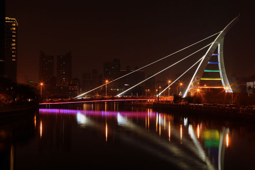 Illuminated mystical bridge at night with vibrant lights reflecting on calm water in an urban setting.