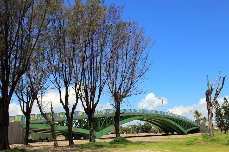 Green mystical bridge with colorful railings surrounded by tall trees under a clear blue sky.