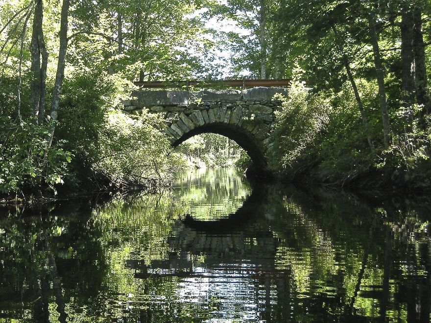 Stone arch mystical bridge over calm water surrounded by lush green trees reflecting another world.