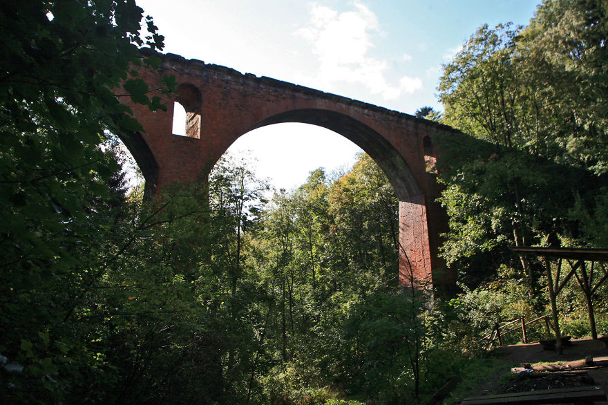 Ancient red brick mystical bridge arching over lush forest under a bright sky, evoking a sense of another world.