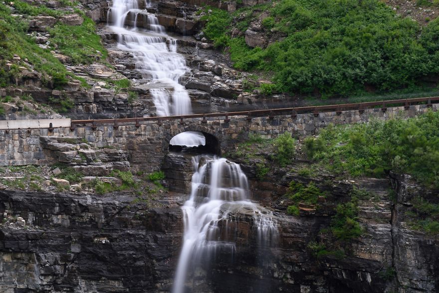 Stone mystical bridge over cascading waterfall surrounded by lush greenery and rocky cliffs in a serene natural setting